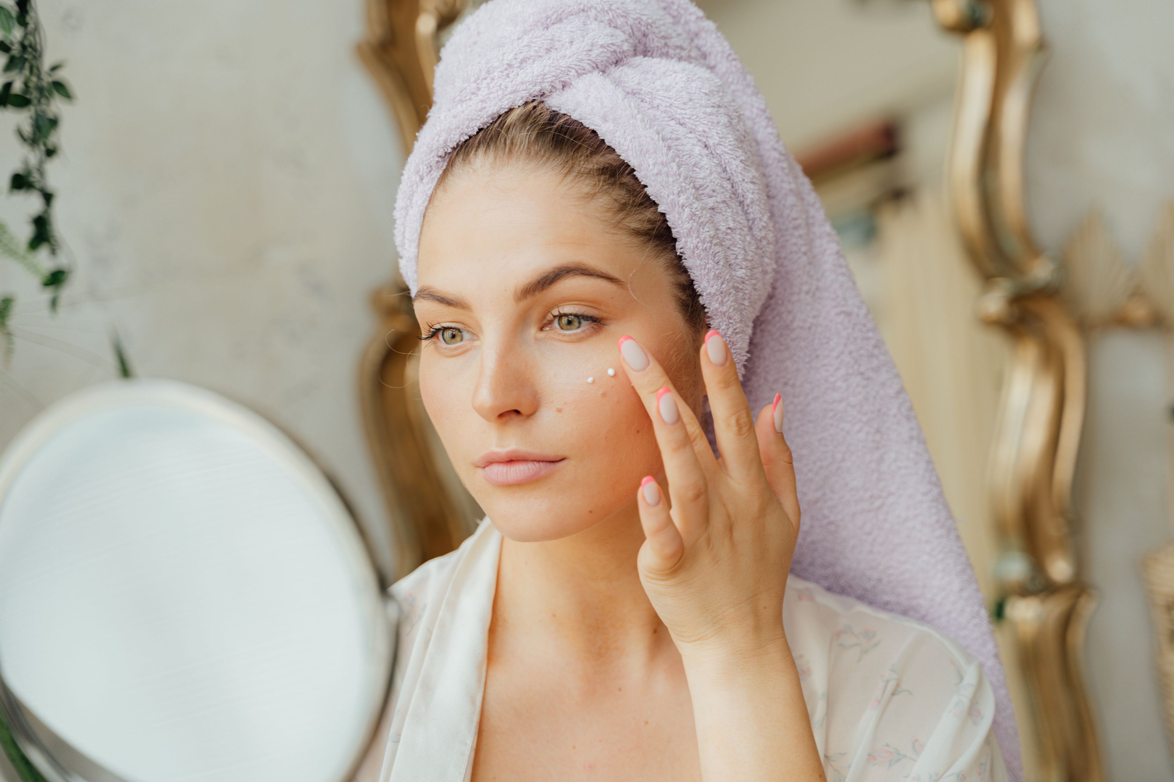 Woman applying cream to her face with a towel on her head, indoors.