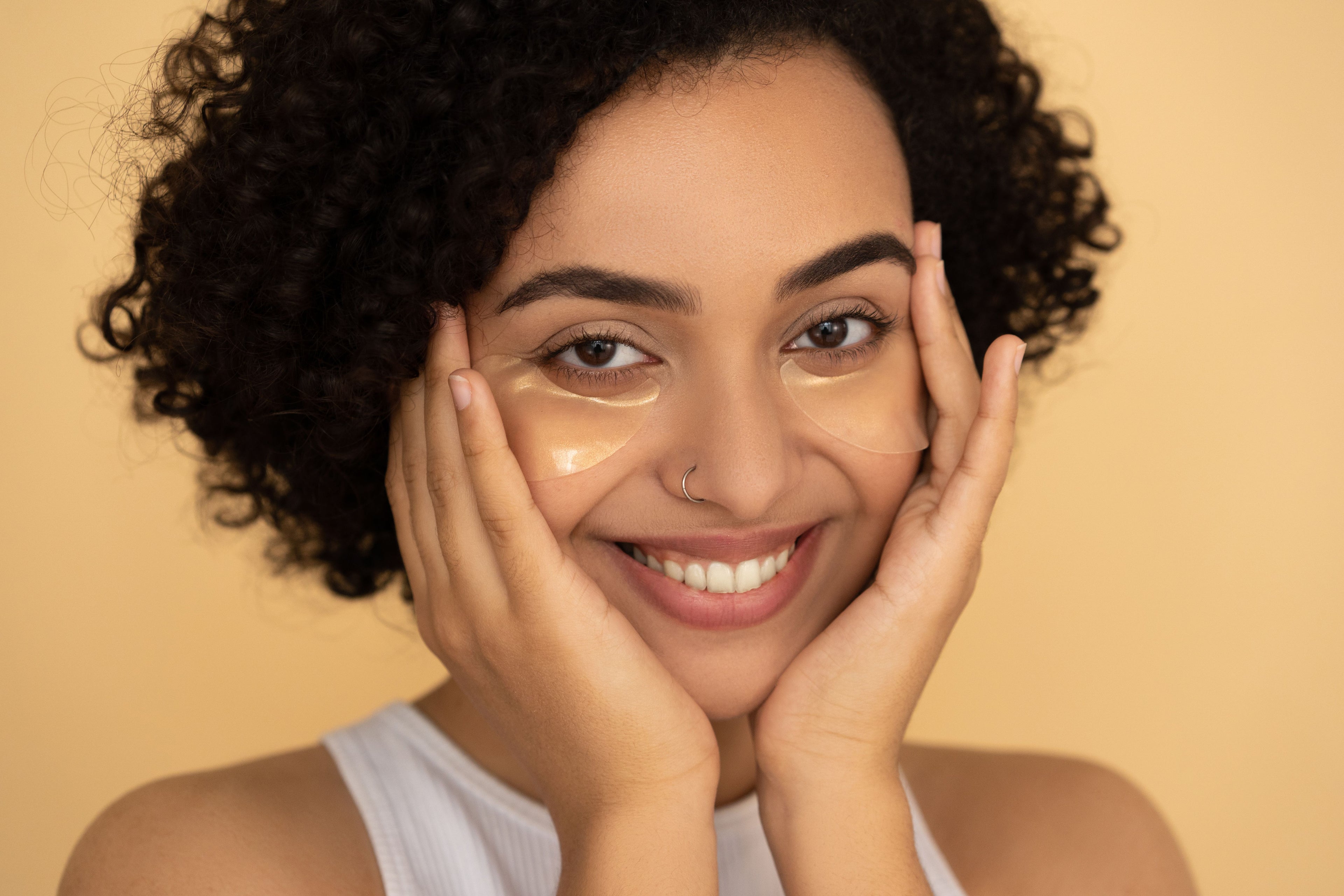 A close-up portrait of a smiling woman with dark curly hair, wearing a clear under-eye concealer or patch on her cheeks, set against a solid warm beige background.