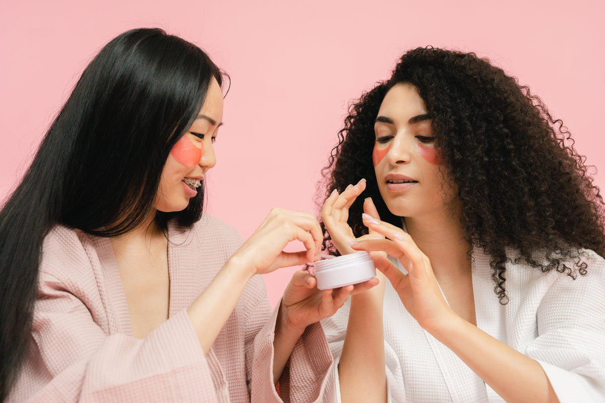 Two women applying cream to their faces against a pink background