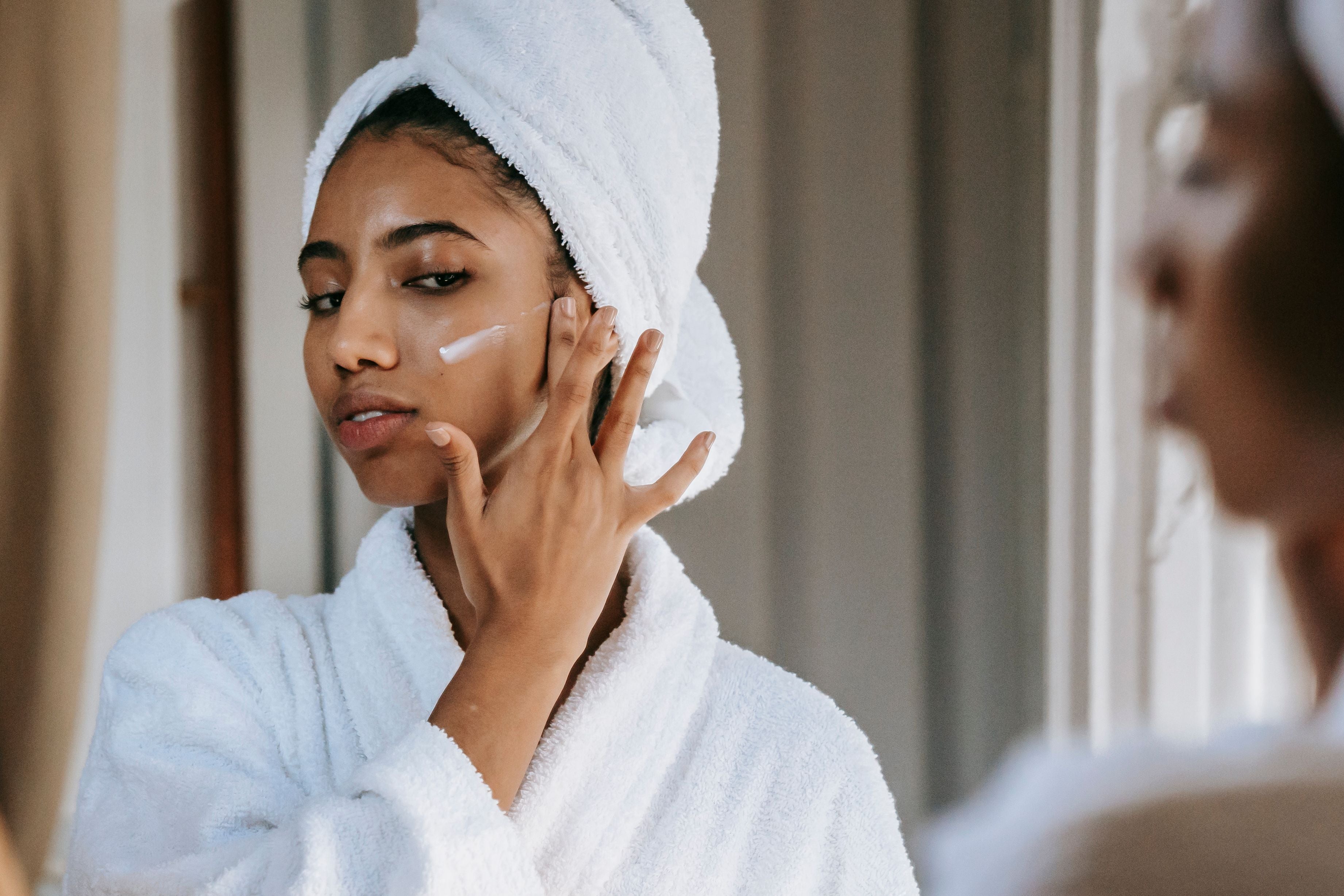 Woman applying cream to her face with a towel on her head in a bathroom setting.